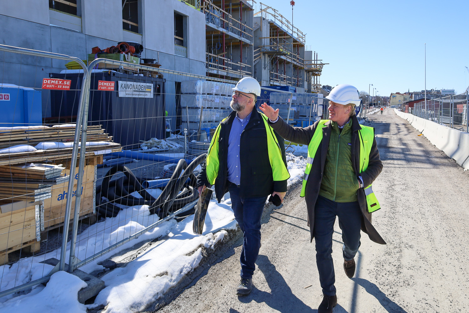 Två män vandrar genom en byggarbetsplats för bostäder. Den ena pekar uppåt fasaden på det blivande flerfamiljshuset